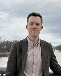 A man stands in front of a river scene in shirt and jacket. 