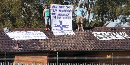 Two men stand on a rooftop holding a sign that reads &quot;Are not we your neighbour don&#039;t mistreat the foreigner&quot;