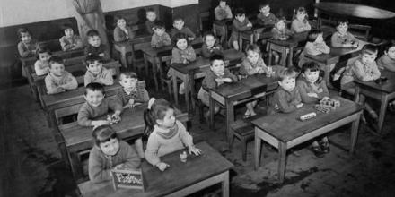 A historic black and white photo of young children sitting together in a classroom.