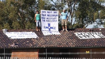 Two men stand on a rooftop holding a sign that reads &quot;Are not we your neighbour don&#039;t mistreat the foreigner&quot;
