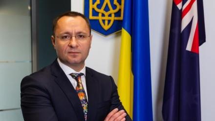 The Ukrainian Ambassador, Vasyl Myroshnychenko, stands with his hands folded. Behind him are the Ukrainian and Australian flags.