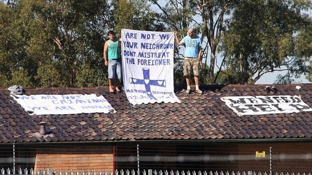 Two men stand on a rooftop holding a sign that reads &quot;Are not we your neighbour don&#039;t mistreat the foreigner&quot;