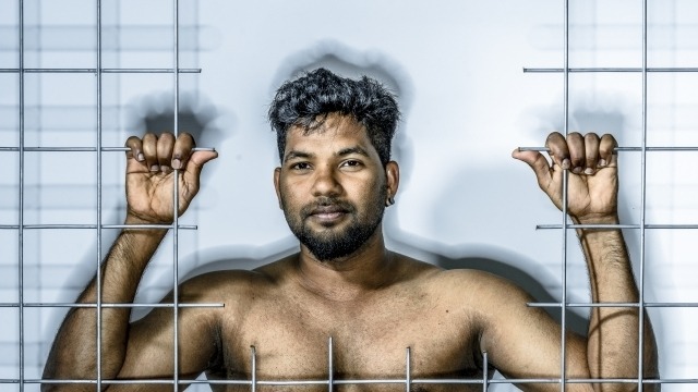 Stories of We Bleed the Same Image of a Tamil refugee, Thanush Selvarasa, standing behind bars.