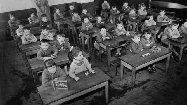 A historic black and white photo of young children sitting together in a classroom.