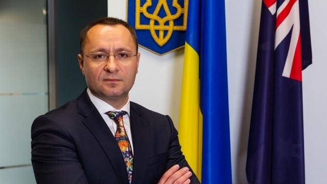 The Ukrainian Ambassador, Vasyl Myroshnychenko, stands with his hands folded. Behind him are the Ukrainian and Australian flags.