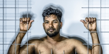 Image of a Tamil refugee, Thanush Selvarasa, standing behind bars.