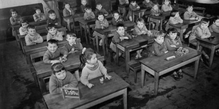 A historic black and white photo of young children sitting together in a classroom.