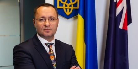 The Ukrainian Ambassador, Vasyl Myroshnychenko, stands with his hands folded. Behind him are the Ukrainian and Australian flags.