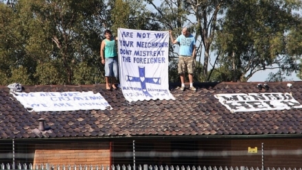 Two men stand on a rooftop holding a sign that reads "Are not we your neighbour don't mistreat the foreigner"