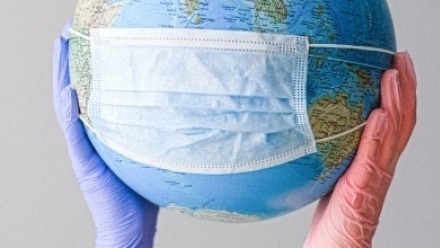 Two gloved hands hold a world globe which has a disposable face mask stretched across it