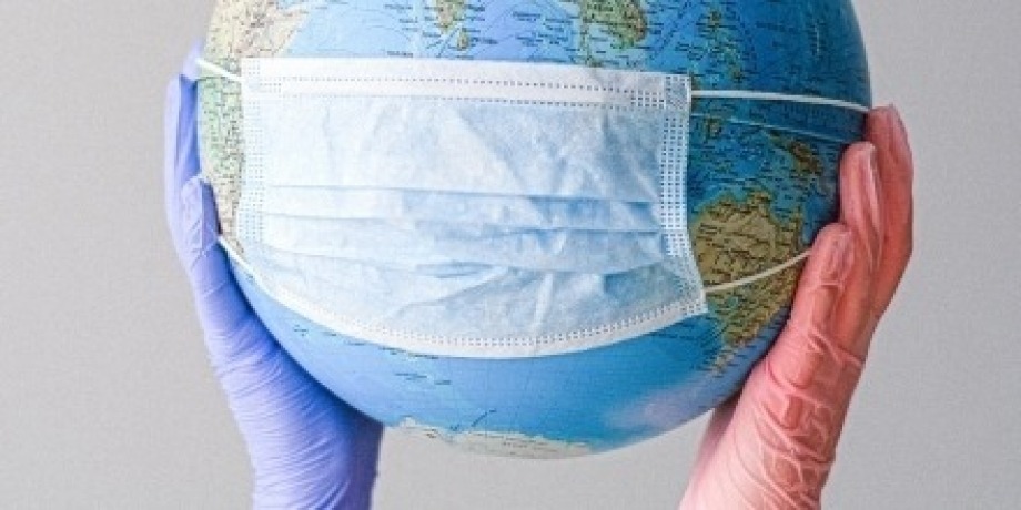 Two gloved hands hold a world globe which has a disposable face mask stretched across it